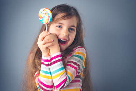 Little kid eating lollipop. Happy beautiful girl with candy isolated on blue background.の写真素材