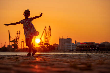 Little girl dancing during sea sunset. Silhouttes of industrial cranes on sea port Varna, Bulgariaの写真素材