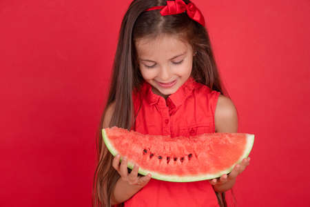 Cute little girl holding, eating  juicy slice of watermelon over red backgroundの写真素材