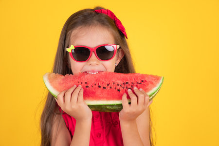 Cute little girl holding, eating  juicy slice of watermelon over yellow backgroundの写真素材