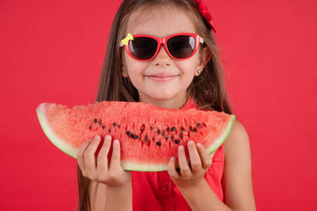 Cute little girl holding, eating  juicy slice of watermelon over red backgroundの写真素材