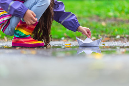 Happy child girl with colorful rubber boots playing with paper boat in puddle in autumn on natureの写真素材