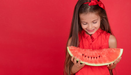 Cute little girl holding, eating  juicy slice of watermelon over red backgroundの写真素材