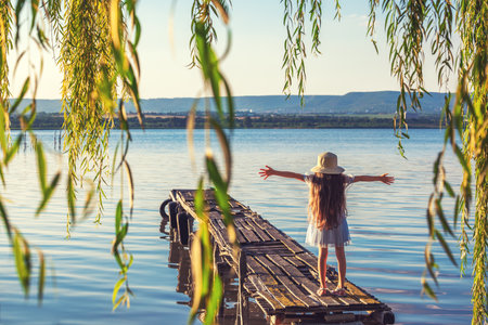 Girl on a old wooden fishing pier and willow tree enjoying beautiful sunset over the sea lakeの写真素材