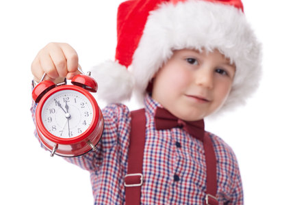 Christmas boy with red alarm clock, smiling little man in red Santa claus hat, tie and suspender posing on white backgroundの写真素材