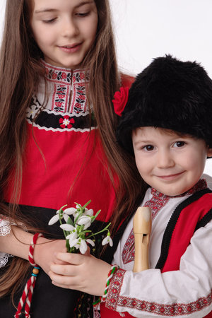 Bulgarian kids boy and girl in traditional folklore costumes with spring flowers snowdrop and handcraft wool bracelet martenitsa symbol of Baba Martaの写真素材