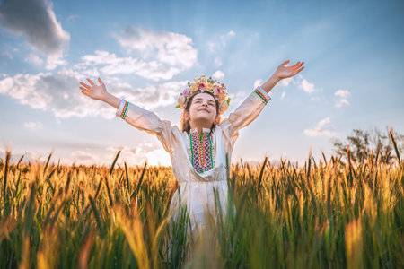 Beautiful young girl with flower chaplet, ethnic folklore dress with traditional Bulgarian embroidery during sunset on a wheat agricultural fieldの写真素材