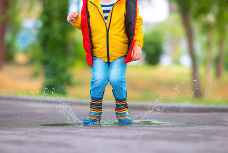 Happy toddler boy with umbrella and color rubber boots jump on a rainy puddle in a park, carefree childhoodの写真素材