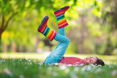 Cute toddler girl in rain rubber boots playing outside in the green park with blooming field of daisy flowers, kid portraitの写真素材