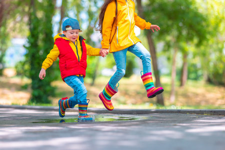 Happy kids girl and boy with umbrella and colorful rubber rain boots playing outdoor and jumping in rainy puddleの写真素材
