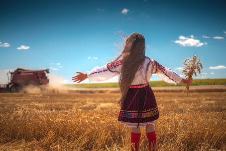 Young girl with traditional Bulgarian folklore costume at the agricultural wheat field during harvest time with industrial combine machineの写真素材