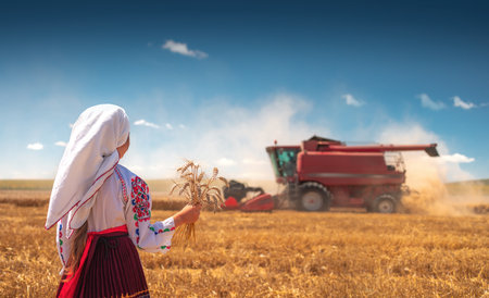 Young girl with traditional Bulgarian folklore costume at the agricultural wheat field during harvest time with industrial combine machineの写真素材