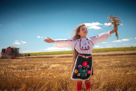 Young girl with traditional Bulgarian folklore costume at the agricultural wheat field during harvest time with industrial combine machineの写真素材