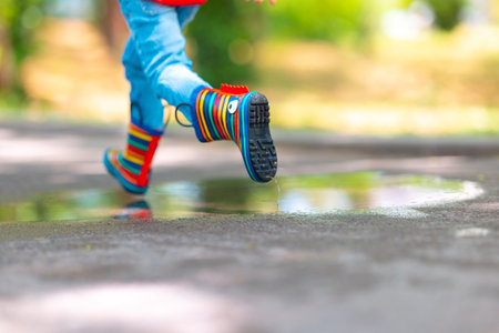 Feet of child in colorful  rubber rain boots jumping over rainy puddle in a park