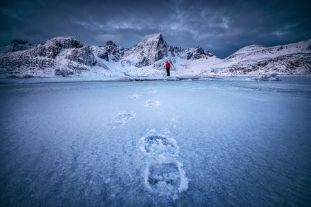 Scandinavian winter Landscape of Snowy Mountain peaks and sea coast, frozen lake water with footsteps in snow and photographer person with camera. Lofoten, Norwayの写真素材