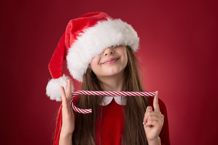 Smiling girl hold Christmas candy cane and wear red Santa Claus hat in red backgroundの写真素材