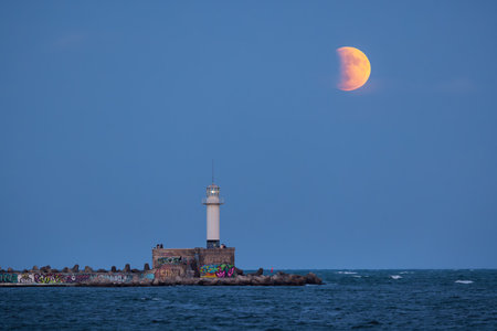 Full moon and lunar eclipse, twilight landscape over sea horizon and lighting lighthouse from the...