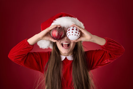 Smiling girl hold Christmas tree balls decoration into his eyes  and wear Santa Claus hat dress portrait in red backgroundの写真素材