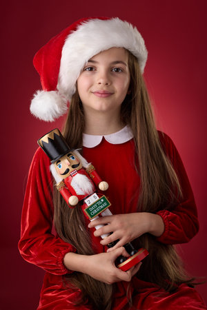 Little girl with red Santa hat holding the wooden Christmasc toy Nutcracker portrait on red...