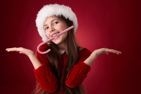 Smiling girl hold Christmas candy cane and wear red Santa Claus hat in red backgroundの写真素材