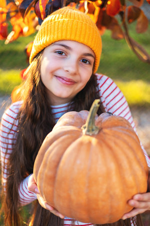Smiling young Girl in Orange Beanie Holds Large Pumpkin in Bright Autumn Garden During Golden Sunset Light and Color fallen tree leavesの写真素材