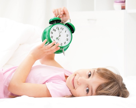 Beautiful little girl with alarm clock on bed in the bedroomの写真素材