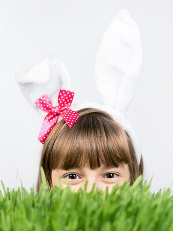 Beautiful little girl dressed in Easter bunny ears peeking from behind the green grass on a gray backgroundの写真素材
