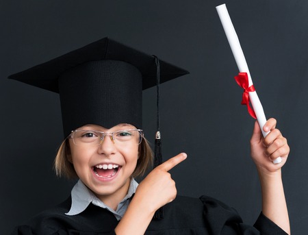 Close-up portrait of happy girl in mortar board holding diploma at the black chalkboard in classroomの写真素材