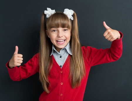 Portrait of adorable young girl showing thumbs up sign using both hands at the black chalkboard in classroom.の写真素材