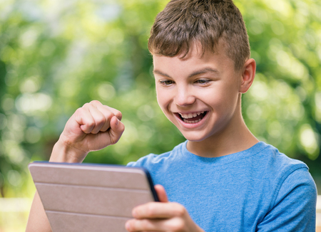 Outdoor portrait of happy teen boy 12-14 year old with tablet pc watching video in summer parkの写真素材