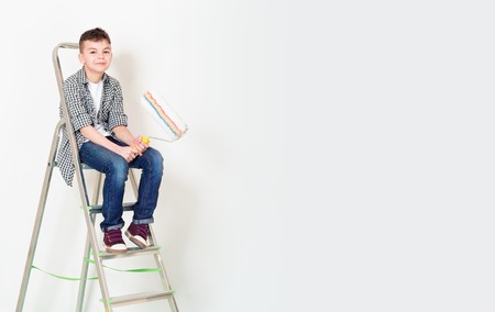 Happy boy makes repairs at home. Smiling teenager with roller sit on a ladder. Wall for text.の写真素材