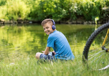 Teen boy with tablet pc and headphones listening to music or watching video in summer park at lakeside. Child resting after biking.の写真素材