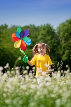 Cute happy little girl playing outdoors with windmill toy against blue summer sky background. Laughing kid in green field. Best childhood concept.の写真素材