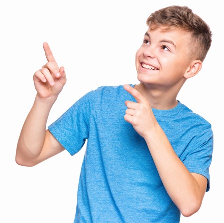 Half-length emotional portrait of caucasian teen boy wearing blue t-shirt. Funny teenager pointing and looking upwards while smiling, isolated on white background. Handsome happy child pointing at copy space.の写真素材