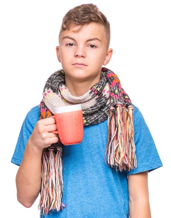 Healthcare and medicine concept - ill teen boy with flu holding cup of hot tea or milk. Unhappy teenager with mug, isolated on white background. Sad child wearing yellow t-shirt and scarf drinks water.の写真素材
