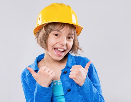 Emotional portrait of attractive girl wearing safety yellow hard hat. Beautiful child making thumbs...