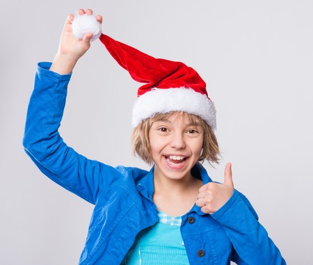 Attractive caucasian little girl wearing Santa Claus red hat. Funny cute smiling child 10 year old thumbs up gesture and looking at camera on gray background. Winter holiday christmas concept.の写真素材