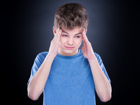Stress and headache - teen boy having migraine pain. Handsome child suffering from a headache. Unhappy caucasian teenager touching his head, with eyes closed, on black background.の写真素材