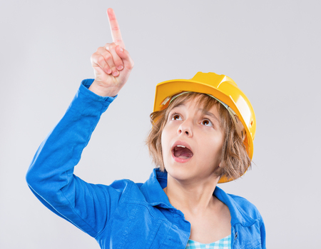 Emotional portrait of caucasian little girl wearing safety yellow hard hat. Shocked or surprised child pointing up and looking away, isolated on white background.の写真素材