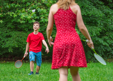 Mother and son playing badminton in meadow with forest in background. Child with badminton rackets in hand. Kid have fun in summer park at day.の写真素材