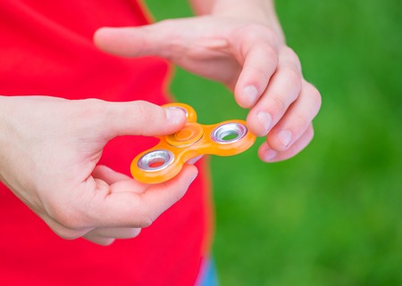 Male child hand holding popular fidget spinner toy - close up, outdoors. Boy playing with a orange Spinner in park.の写真素材