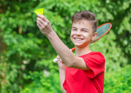 Young teen boy playing badminton in meadow with forest in background. Child with badminton rackets in hand. Kid have fun in summer park at day.の写真素材