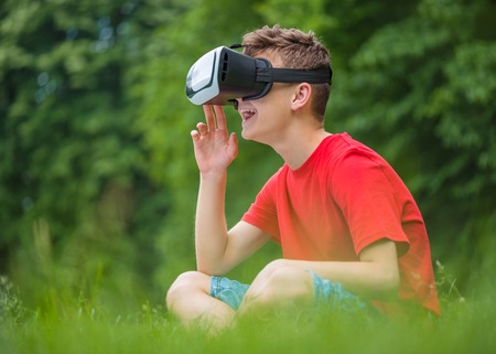 Playful teen boy using virtual reality goggles outdoor in summer park. Teenager looking in VR...