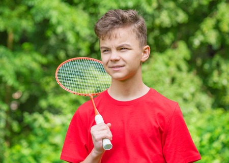 Young teen boy playing badminton in meadow with forest in background. Child with badminton rackets in hand. Kid have fun in summer park at day.の写真素材