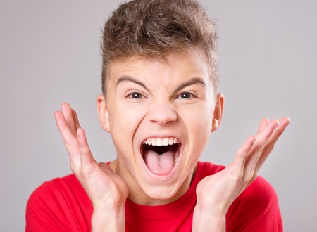 Emotional portrait of irritated shouting teen boy. Furious teenager screaming and looking with anger at camera. Handsome outraged child shouting out loud on gray background.の写真素材