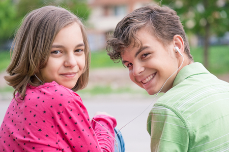 Happy teenage boy and girl with headphones are using gadget, talking and smiling while sitting on the stairs outdoors. Young sister and brother teens playing on mobile phone and listening to music.の写真素材