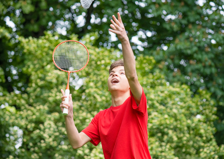 Young teen boy playing badminton in meadow with forest in background. Child with badminton rackets in hand. Kid have fun in summer park at day.の写真素材
