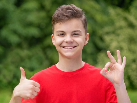 Caucasian smiling teen boy making thumbs up and ok gesture. Handsome funny teenager wearing red t-shirt, at summer park. Happy child looking at camera - outdoors portrait.の写真素材