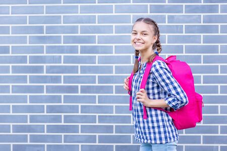 Beautiful student teenager schoolchild with backpack looking at camera. Smiling cute child with bag. Teen girl with braided hair against a brick wall outdoors. Childhood and Back to school concept.の写真素材