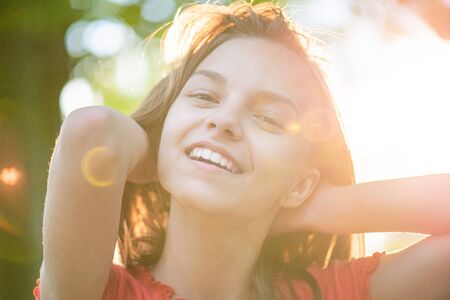 Close up portrait of teenage girl with sun rays filtering through her hair. Happy smiling teen at summer park in flare sunshine. Child looking at camera during sunset.の写真素材